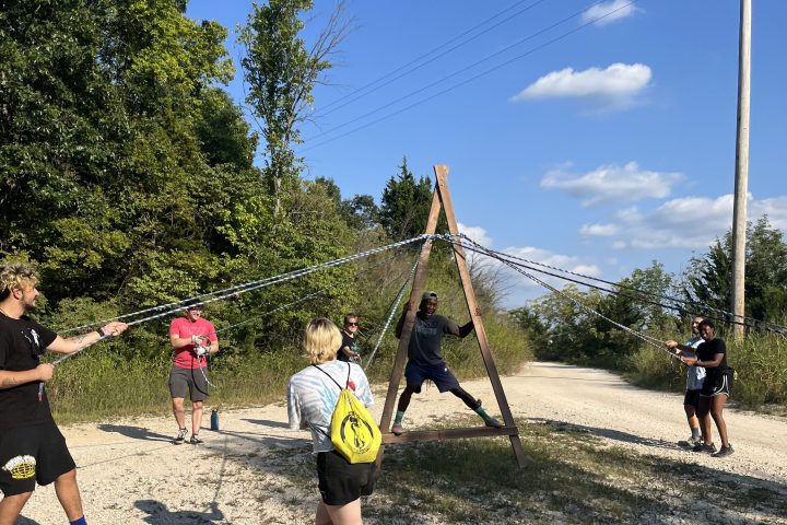 A team Building group on a gravel clearing participating in a team building challenge, the Merry A Frame.