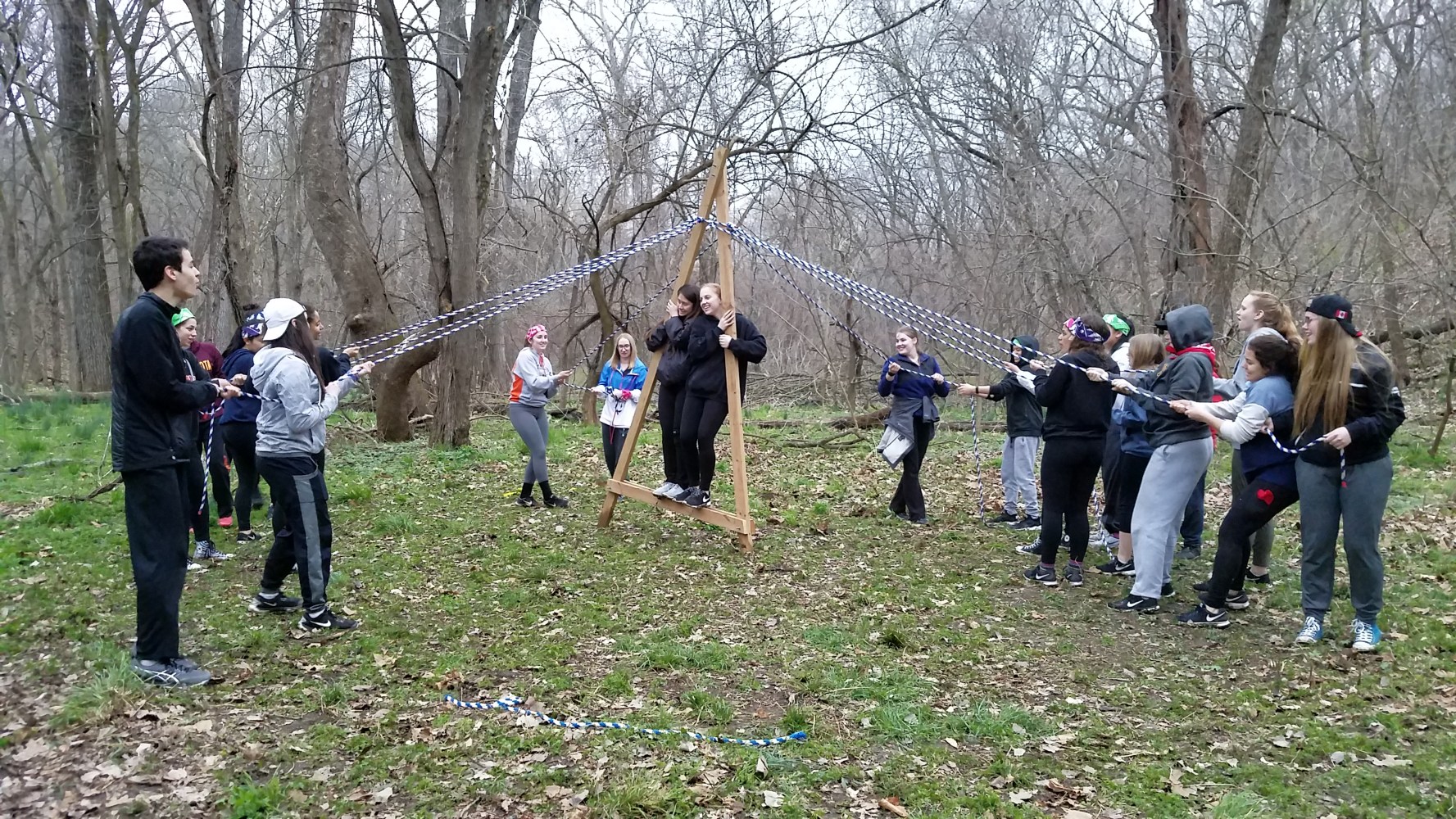 Group outdoors holding ropes supporting a person standing on a wooden structure.