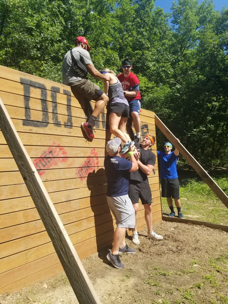Group helping a person climb over a wooden wall in an outdoor obstacle course.
