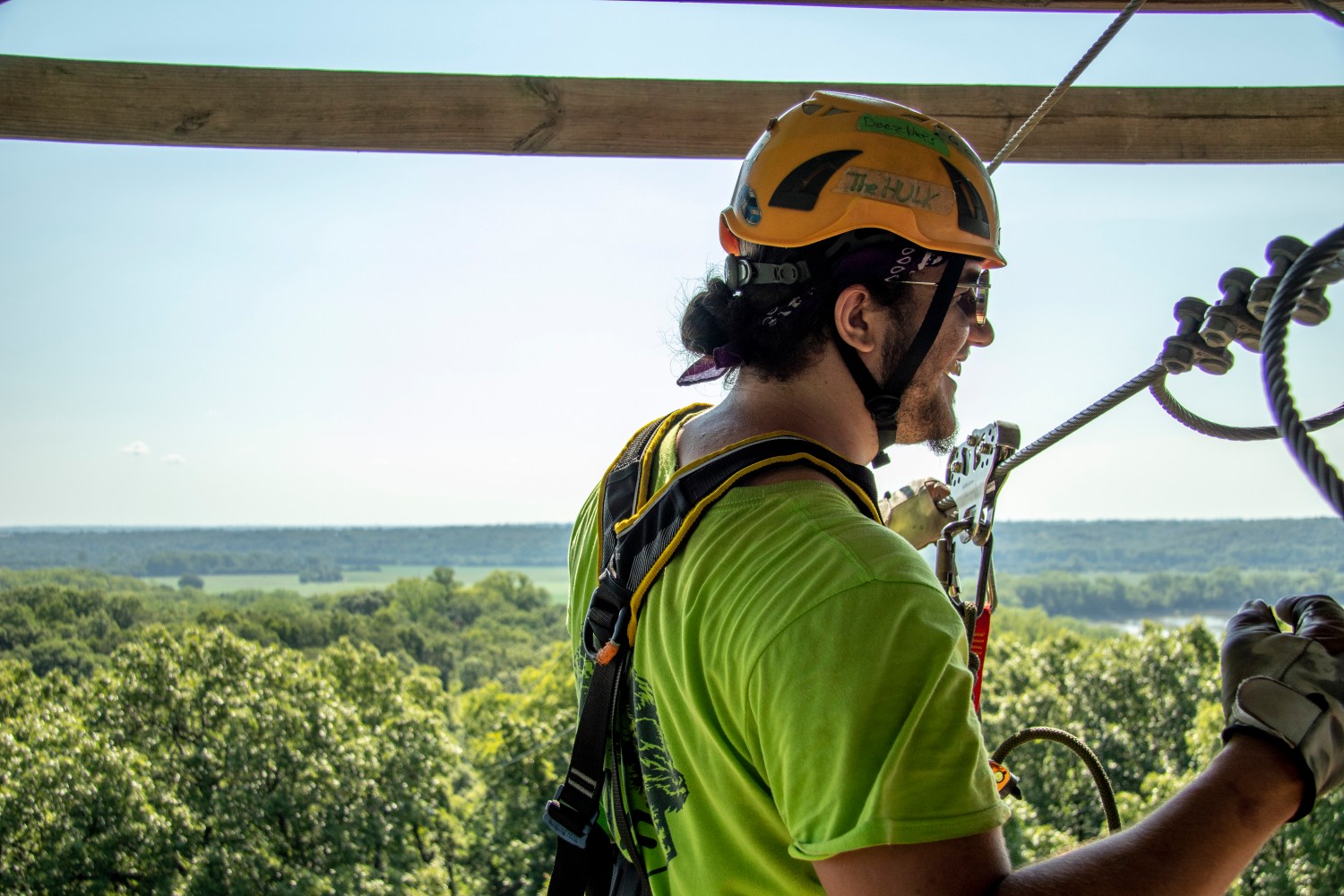 Person in safety gear harnessed, smiling while preparing for zipline, overlooking a wooded landscape.