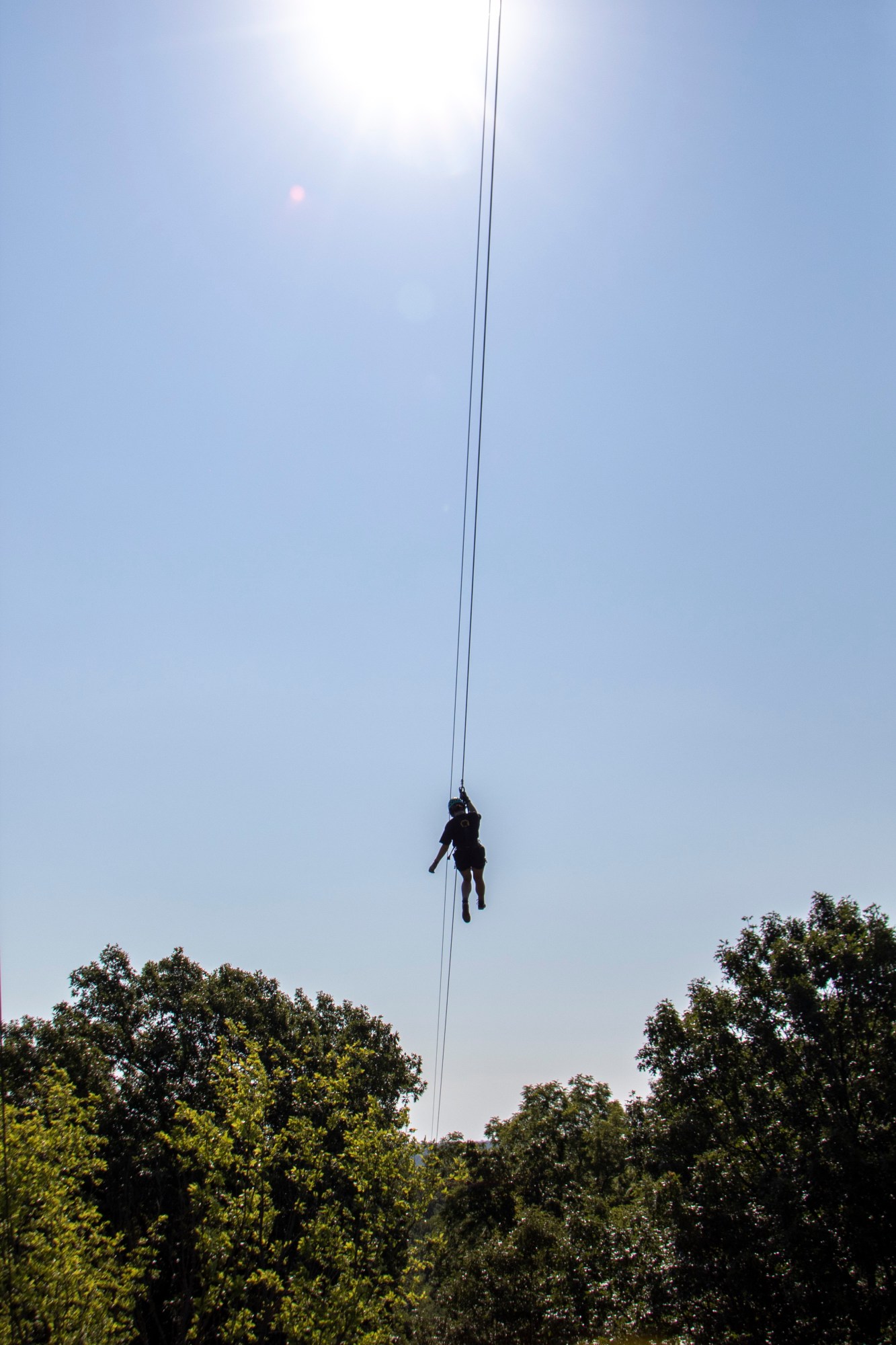 Silhouette of a person hanging from a cable against a bright sunlit sky above trees.